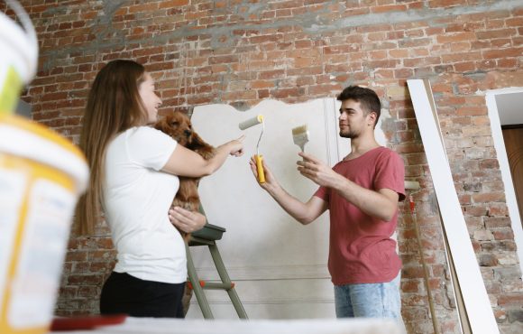 Young couple doing apartment repair together themselves. Married man and woman doing home makeover or renovation. Concept of relations, family, pet, love. Painting the wall, holding the dog.
